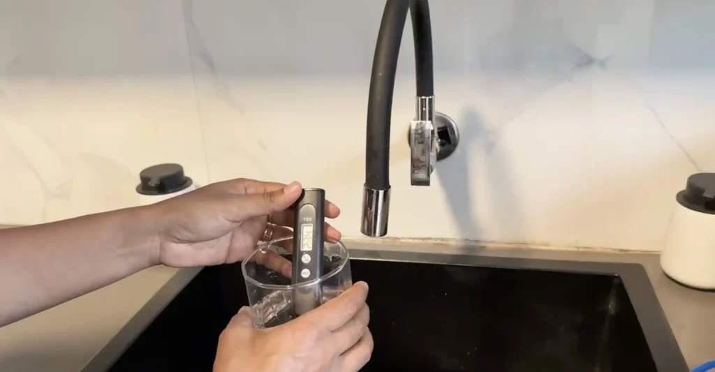 A person using a handheld TDS meter to measure the dissolved solids level in a glass of drinking water in a home kitchen.