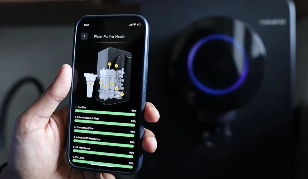 Smartphone displaying a water purifier app with real-time filter health status, component life, and maintenance information while the purifier is visible in the background.